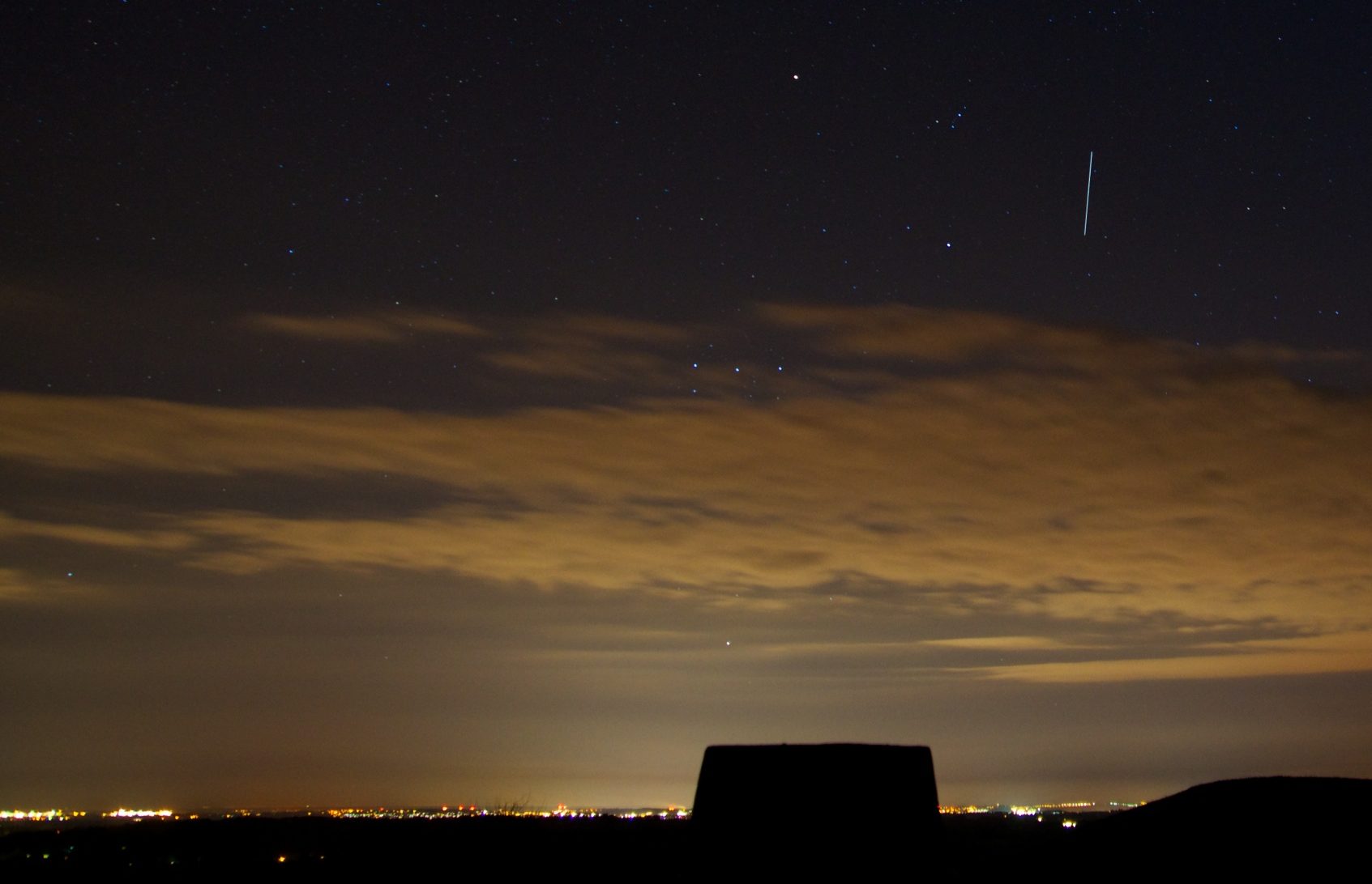Long exposure image taken from Old Winchester Hill in Hampshire on a fairly clear night, showing a vertical streak of light, which marks the passage of the International Space Station. City lights fill the horizon, and in the foreground is the silhouette of a trig point obelisk.