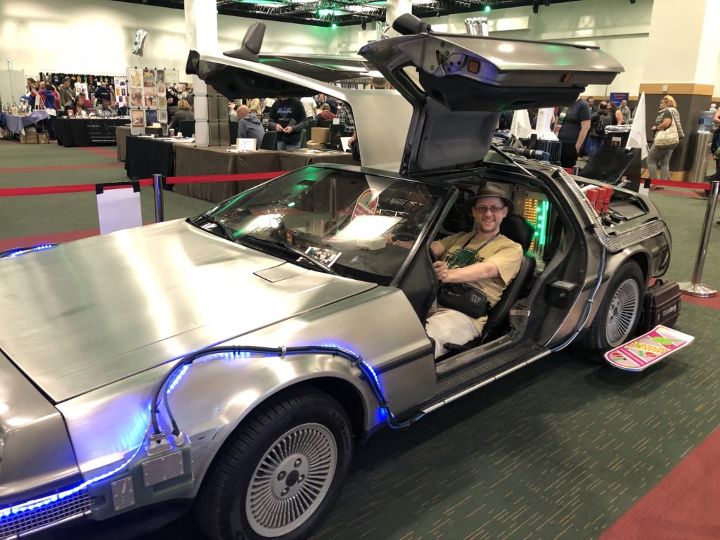 Me sitting at the wheel of a Back To The Future themed DeLorean on the exhibition floor of WorldCon. I'm grinning (as you'd expect) and a hoverboard appears to hover next to the rear wheel.