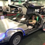 Me sitting at the wheel of a Back To The Future themed DeLorean on the exhibition floor of WorldCon. I'm grinning (as you'd expect) and a hoverboard appears to hover next to the rear wheel.