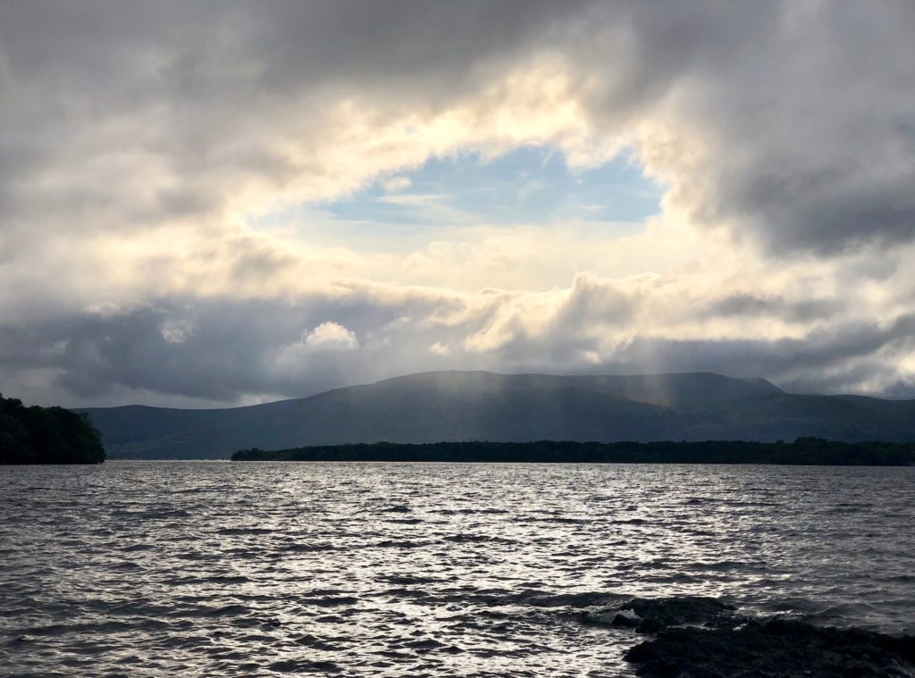 Blue sky peeping through a fortuitously mountain-shaped gap in the clouds above a mountain range, as seen across the choppy waters of Loch Lomond.