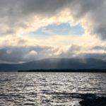 Blue sky peeping through a fortuitously mountain-shaped gap in the clouds above a mountain range, as seen across the choppy waters of Loch Lomond.