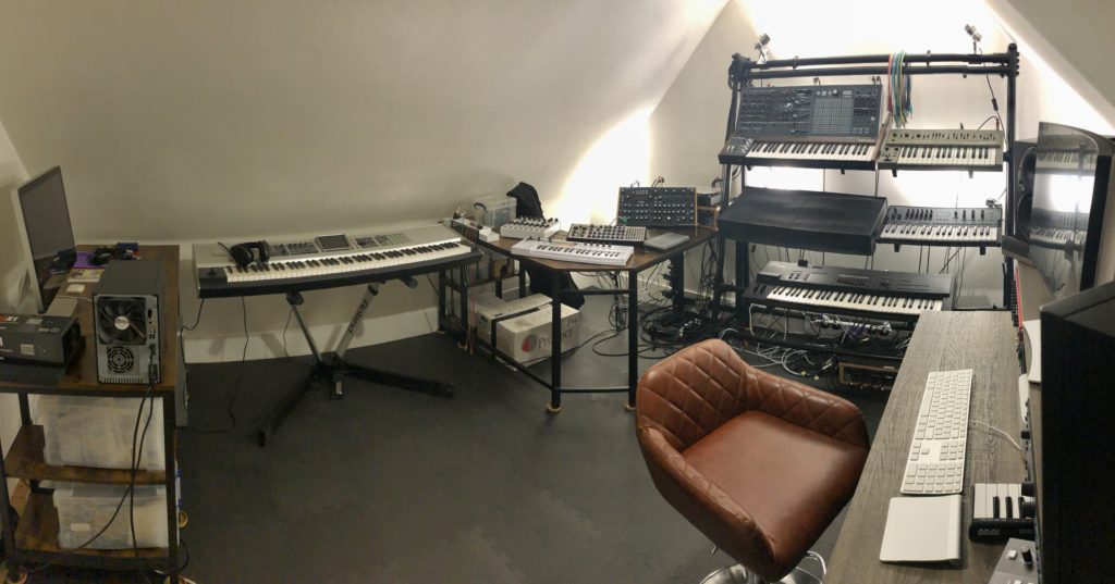 Wide angle shot of my music studio under the sloping roof of my house. Featuring a comfy stool on the right next to the work desk, a stand full of keyboards at the back, various other keyboards and equipment in the corner, etc.