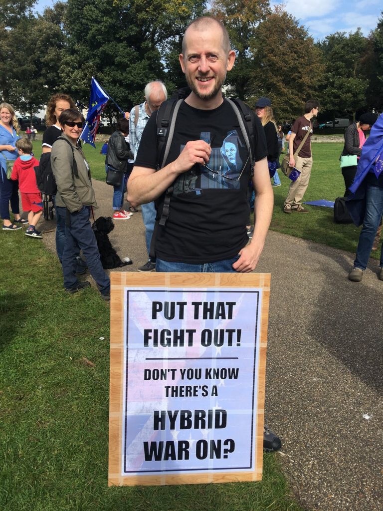 Me standing in a park during the initial gathering of an anti Brexit march. I have a home made poster stuck to a wooden board, reading "Put that fight out! Don't you know there's a hybrid war on?"