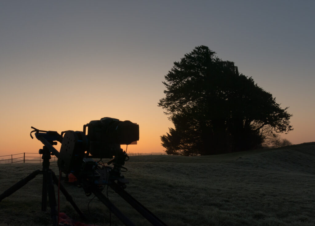 My time lapse tracker rig with camera attached, shown at Knowlton Church in Dorset, shooting a time lapse just before sunrise, in a frosty field. Behind the gear is a tree and a glowing orange horizon