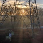 On a small patch of semi-landscaped shrubbery in the middle of a car park is the base of a large electricity pylon. A rising sun casts glory rays towards the observer, streaming down through the metal struts